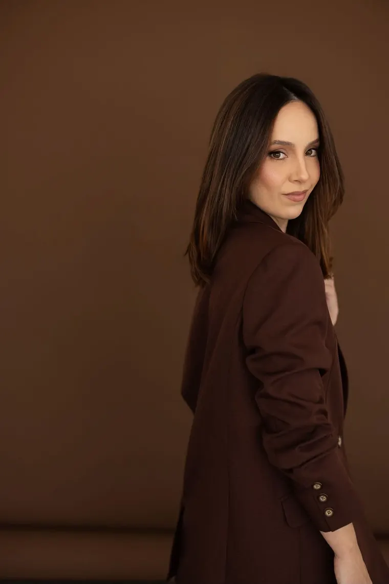 Mari Benante de costas, de blazer marrom, olhando por cima do ombro com um sorriso discreto.