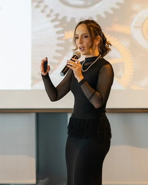 Mari Benante de vestido preto, falando ao microfone em um auditório.